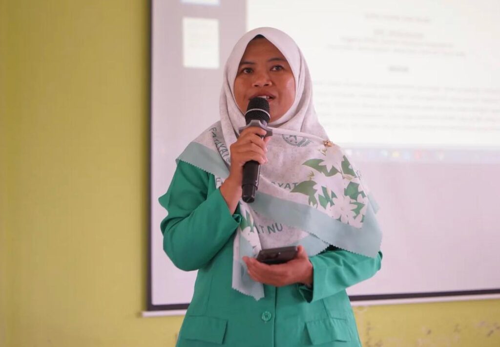 Woman in a green blazer and hijab speaks into a microphone while holding a smartphone presenting in front of a projector